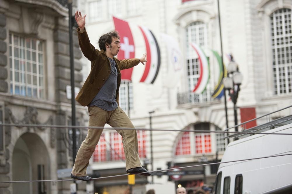 a tightrope walker in a brown trousers and jacket walks on an outdoor highwire against a backdrop of grand buildings and international flags
