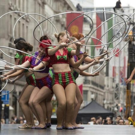 Seven performers in colourful outfits spin hula hoops on an outdoor stage, while an eighth performer in a blue top and black shorts observes nearby.