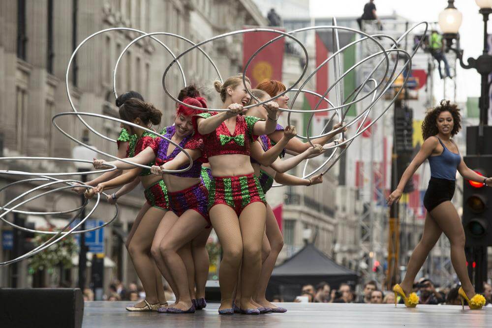Seven performers in colourful outfits spin hula hoops on an outdoor stage, while an eighth performer in a blue top and black shorts observes nearby.