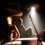 a performer in a red shirt and brown trousers does an upside down headstand, they are spotlit from the side and another performer sits on the floor behind, watching on