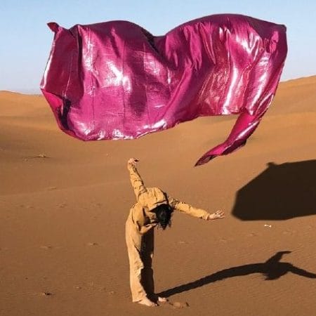 A performer dressed in brown, stands on a sand dune with a large rectangle piece of purple material flying freely above them against the blue sky, both performer and material cast a shadow on the sand. Circostrada
