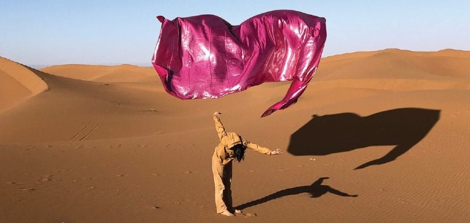 A performer dressed in brown, stands on a sand dune with a large rectangle piece of purple material flying freely above them against the blue sky, both performer and material cast a shadow on the sand. Circostrada