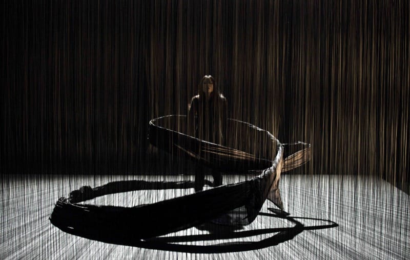 a darkened theatre stade with a performer in the middle of a spiral, of vertical poles physical visual UK theatre