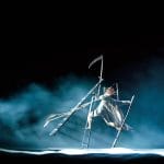 A performer in a flowing white dress balances on stilts amid fog and dramatic lighting, holding long, intricate props.