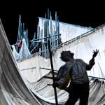 A dramatic theatrical scene with a man holding a staff and gesturing upwards, against an indoor stage backdrop of spotlit tattered sails. Raoul James Thierrée