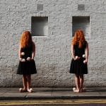 Two red-haired women in black dresses standing barefoot against a white brick wall, each holding juggling props in their hands.