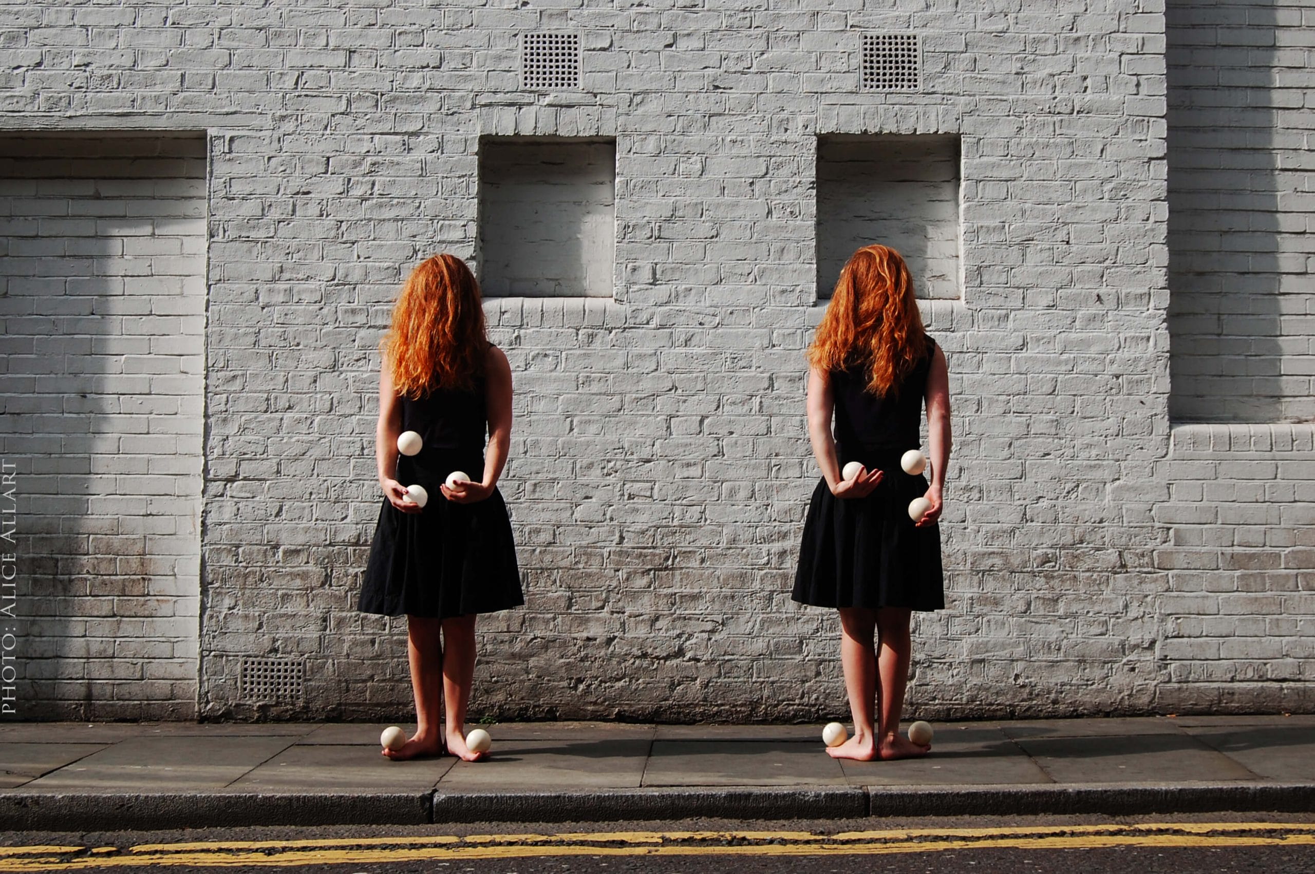 Two red-haired women in black dresses standing barefoot against a white brick wall, each holding juggling props in their hands.