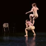 Two dancers performing, one leaping above the other, set against a dark stage with two chairs visible in the background.