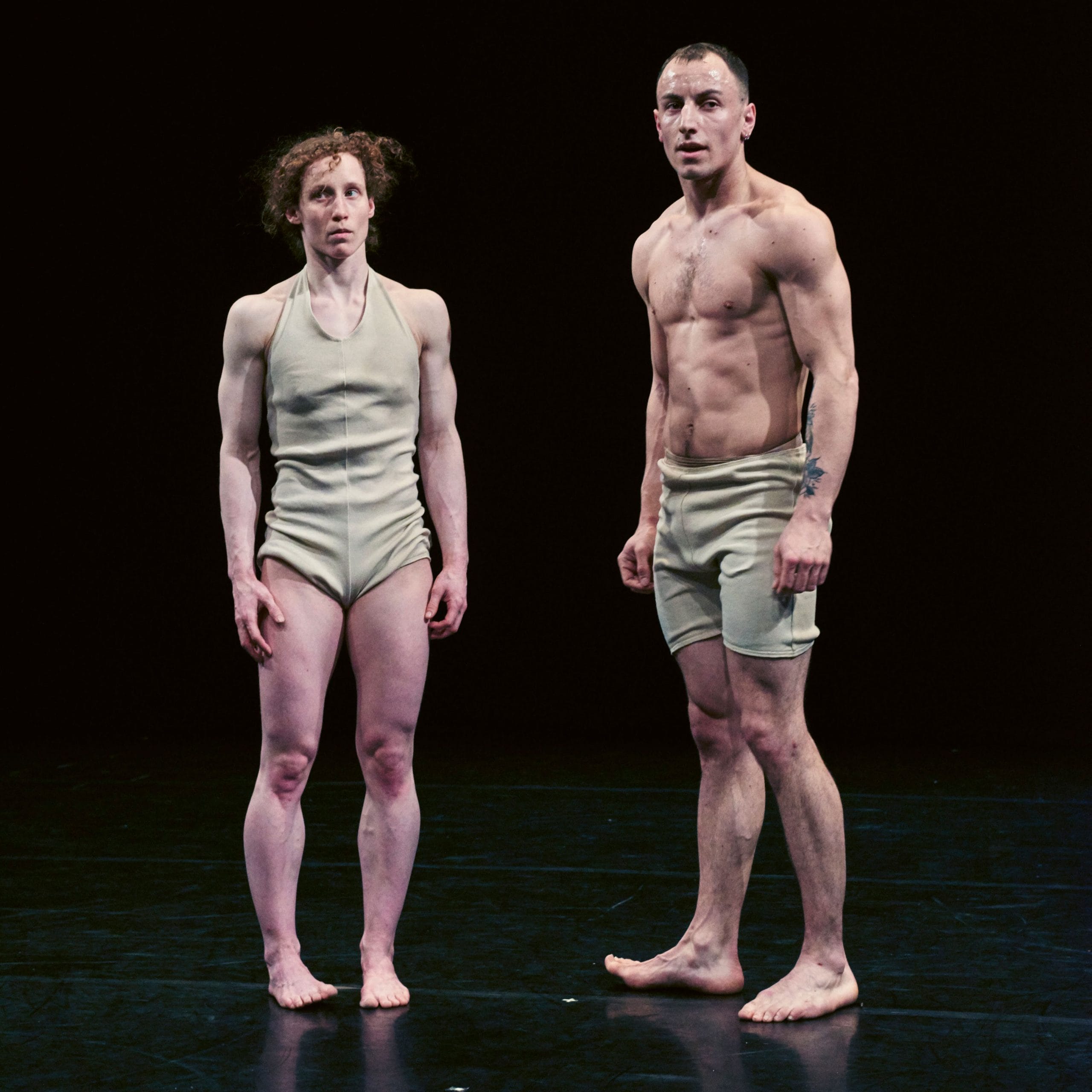 Two performers in fitted flesh-coloured attire standing on a dark stage.