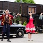 Two people in festive attire hold hands, dancing in front of a vintage car and caravan.