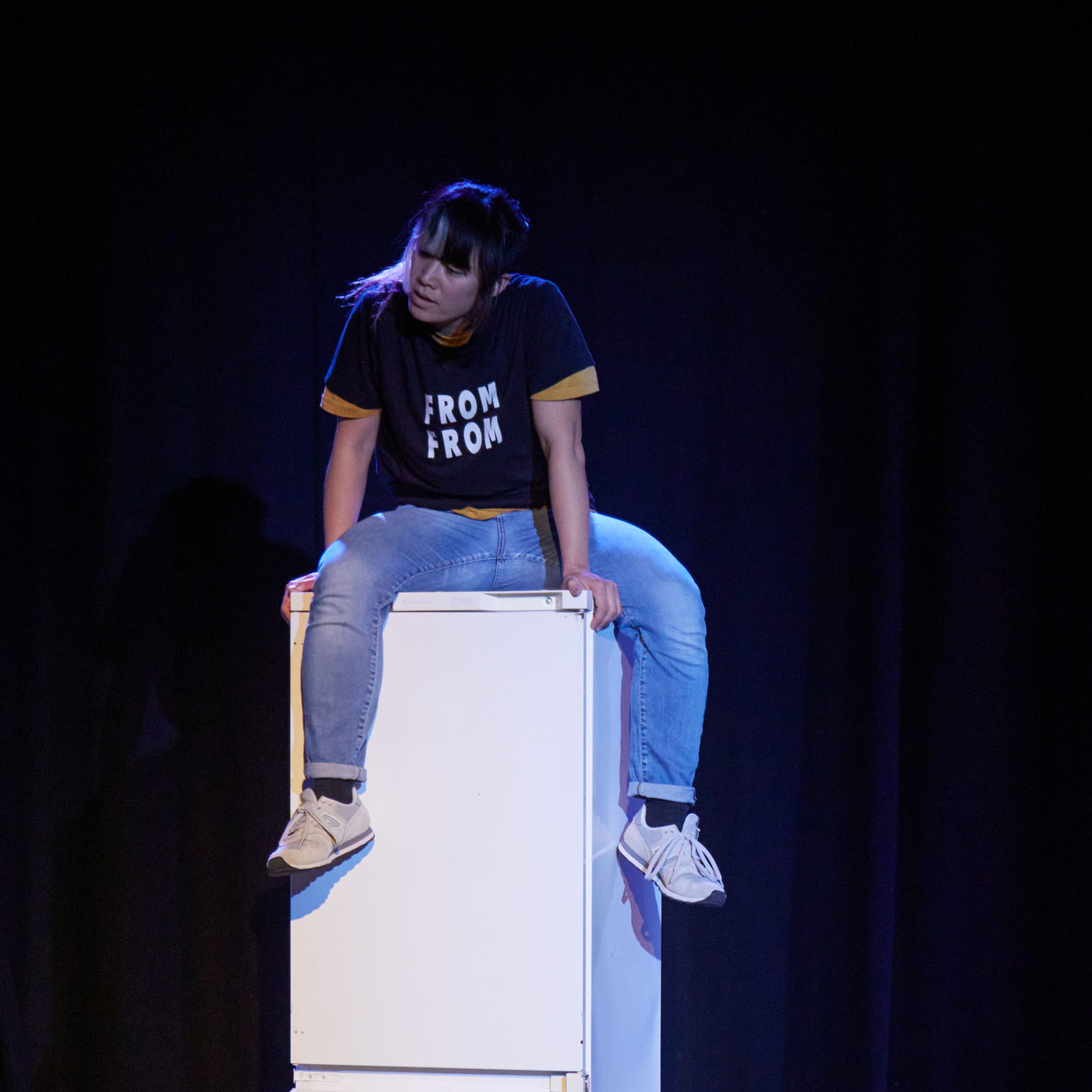East Asian non-binary performer, wearing black t-shirt with white words: From From, with blue jeans, and grey trainers. They are sitting on top of a fridge, holding a strong Krump stance in their body, feeling powerful and frustrated.