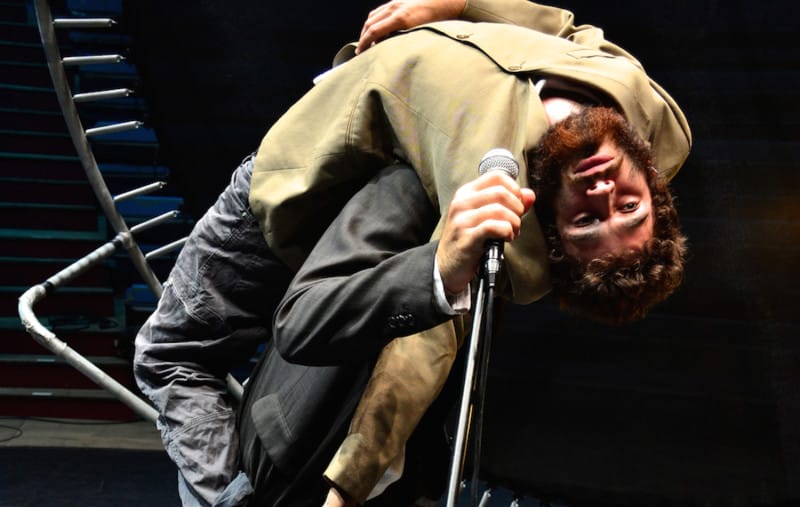A performers with a beard and wearing a green jacket and jeans is carried on the back of a second performer who holds up a microphone to the person they are carrying PASS European contemporary circus