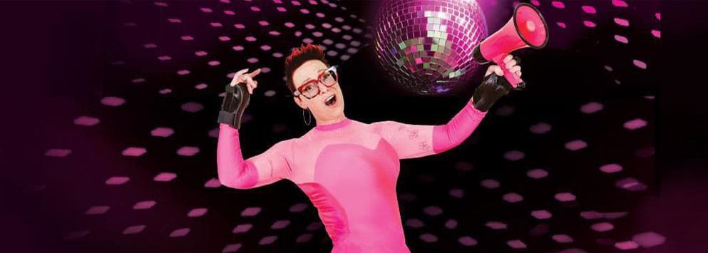 Performer in a pink bodysuit holding a pink loud hailer wears pink framed glasses with short dark hair with pink tips with a glitter ball behind