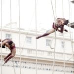 two colourfully dressed aerial performers equipped with safety harnesses performing on a high structure fastened with ropes in front of grand architecture