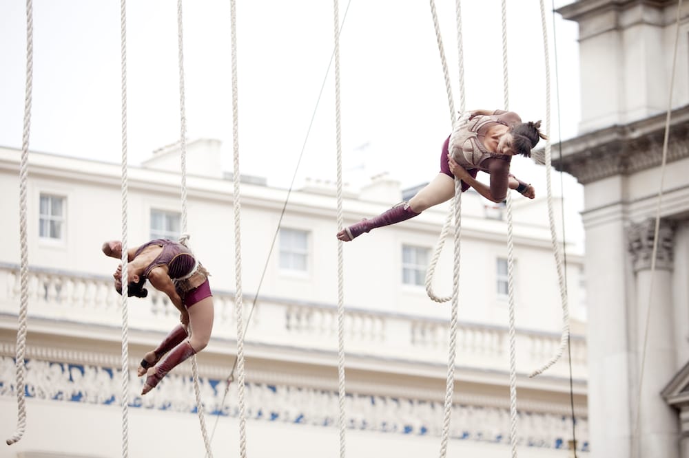 two colourfully dressed aerial performers equipped with safety harnesses performing on a high structure fastened with ropes in front of grand architecture