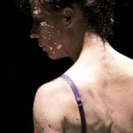 close up of a performer's head and back, wearing a purple bra, with dark hair tied up, they're on a dark stage and have bits of egg shell stuck to their cheek