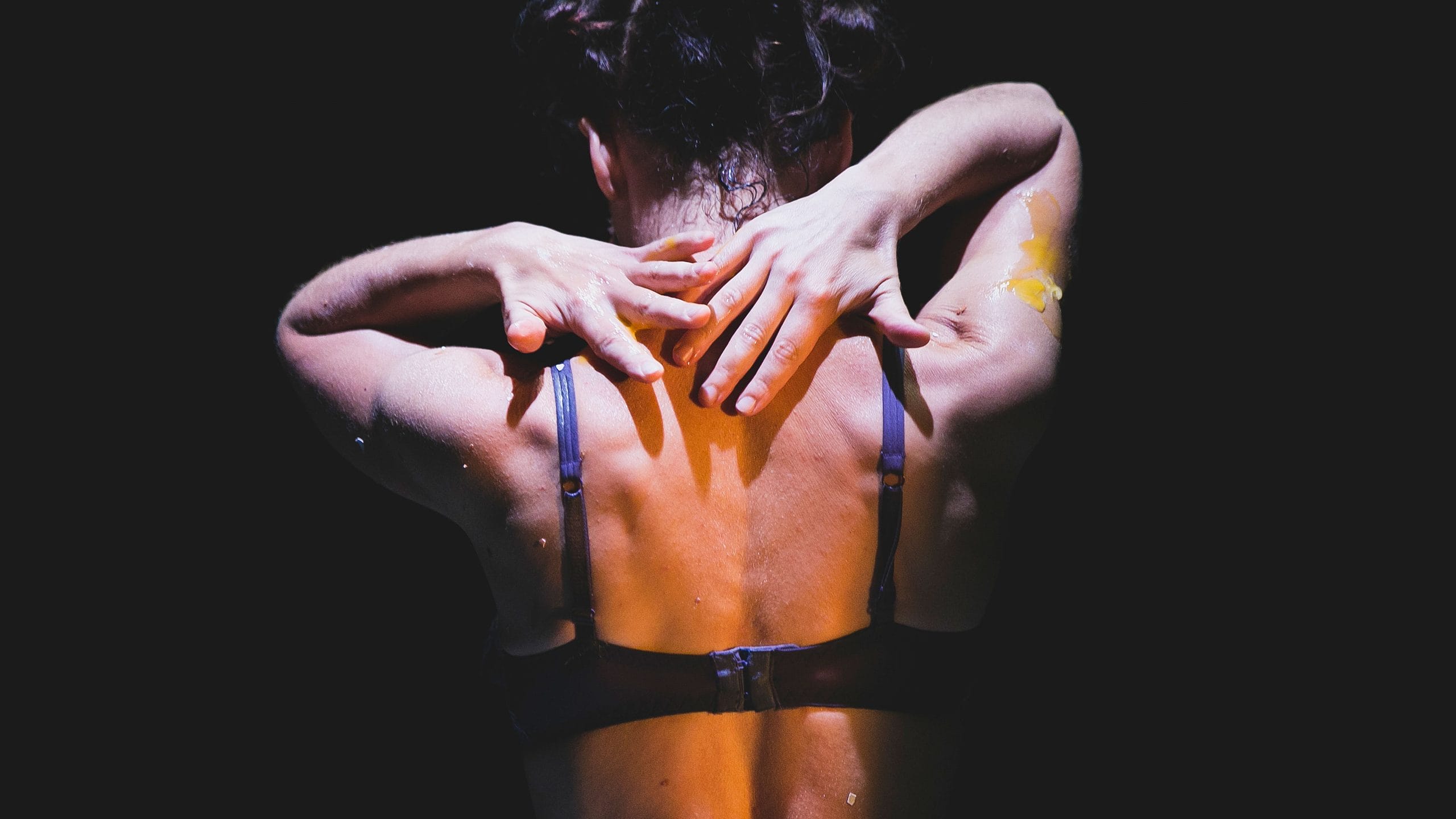 close up of a performer's head and back, wearing a purple bra, with dark hair tied up, they're on a dark stage and on one arm there is some smeared egg yolk