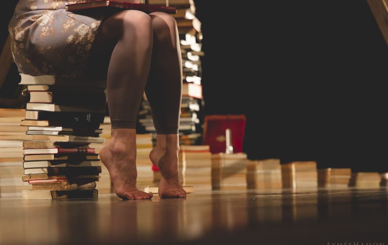 A woman with bare feet sits on a stack of books, holding a closed book in her lap, surrounded by more books in a dimly lit room.