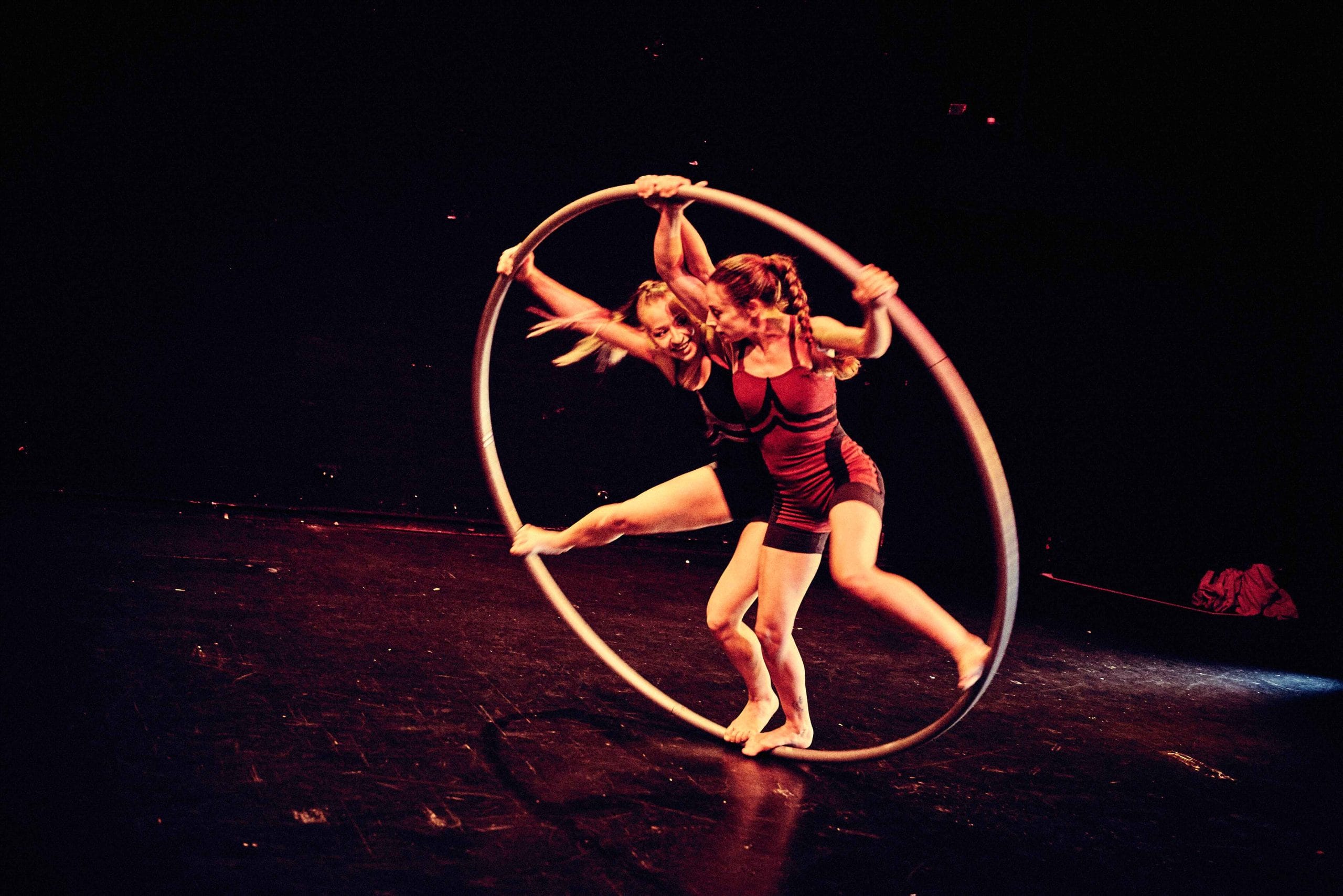 Two acrobatic performers balance within a large hoop on a dimly lit stage. Both are dressed in matching athletic outfits, and the dramatic lighting enhances their movements and expressions.