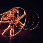 Three acrobats intertwine their movements inside a large hoop under warm, golden stage lighting, creating visually striking moment.