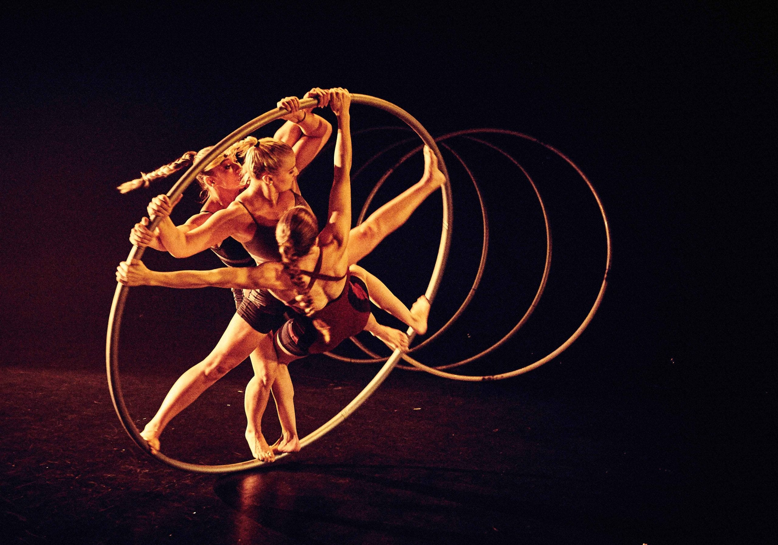 Three acrobats intertwine their movements inside a large hoop under warm, golden stage lighting, creating visually striking moment.