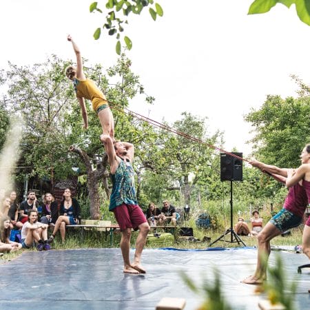 A performer stands balanced on another person's shoulder, reaching upward, while other performers pull a rope to stabilise them. The scene takes place outdoors in a garden with an audience seated around.