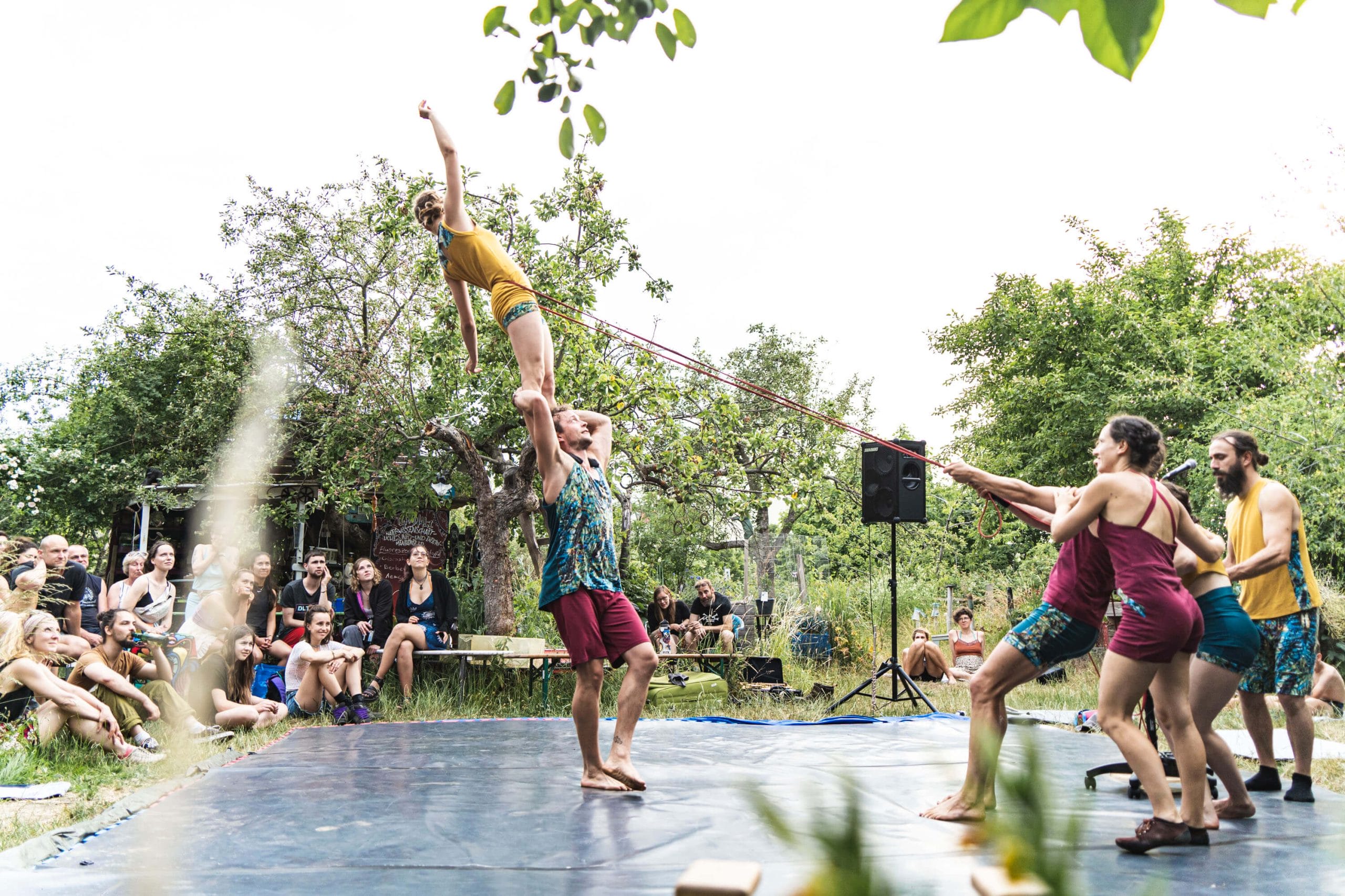 A performer stands balanced on another person's shoulder, reaching upward, while other performers pull a rope to stabilise them. The scene takes place outdoors in a garden with an audience seated around.