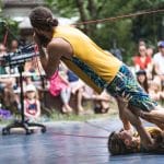 An outdoor performance featuring two people lying down as another performer bends over them, with the audience watching in the background.