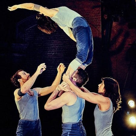three performers on an indoor stage with a brick wall behind and are spotlit, all wear short blue jeans and pale blue vests, a fourth stands on the shoulders of one and is supported by their legs, as they bend backwards preparing to fall or spring back