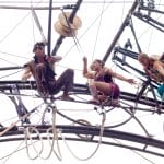 three colourfully dressed performers equipped with safety harnesses navigating obstacles on a high circular wheel-like structure fastened on with ropes
