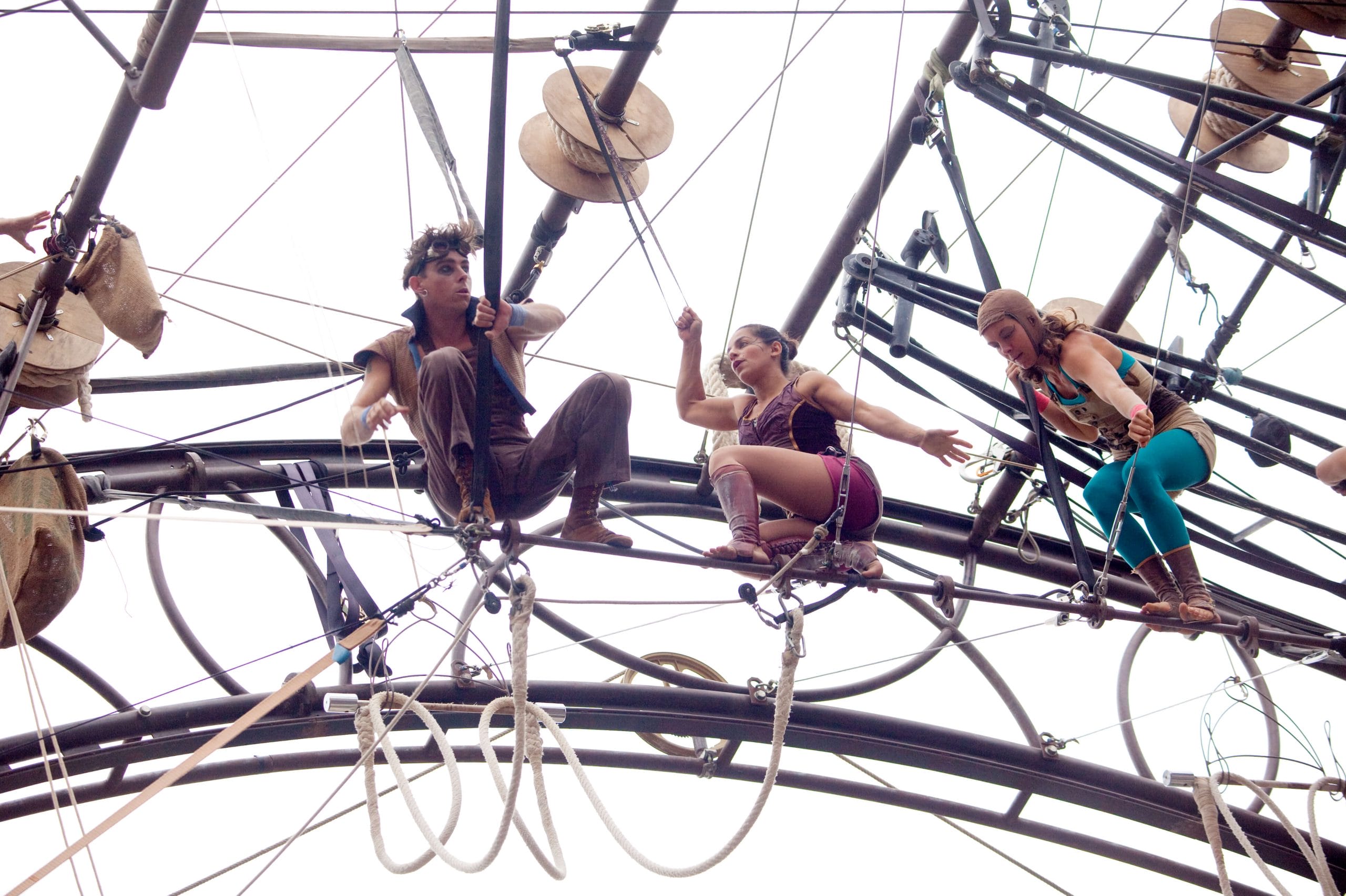 three colourfully dressed performers equipped with safety harnesses navigating obstacles on a high circular wheel-like structure fastened on with ropes