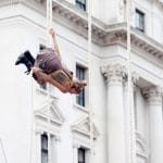 a person with blonde tied up hair and a pale brown outfit swinging on a rope in front of a white historic building, creating a captivating visual scene