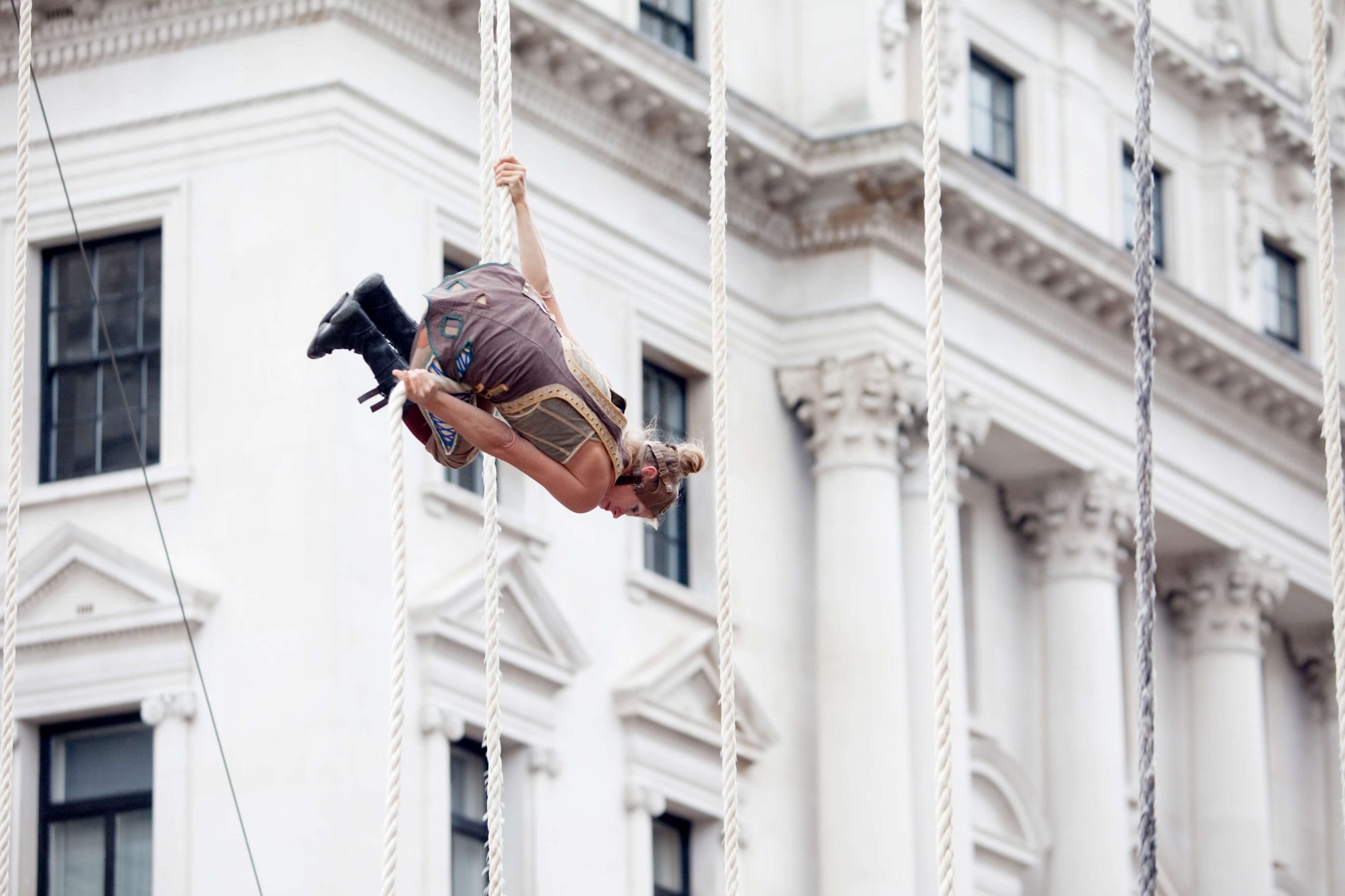 a person with blonde tied up hair and a pale brown outfit swinging on a rope in front of a white historic building, creating a captivating visual scene