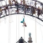 a group of aerial performers hanging from a circular rig above a draped statue, showcasing a daring and dynamic display