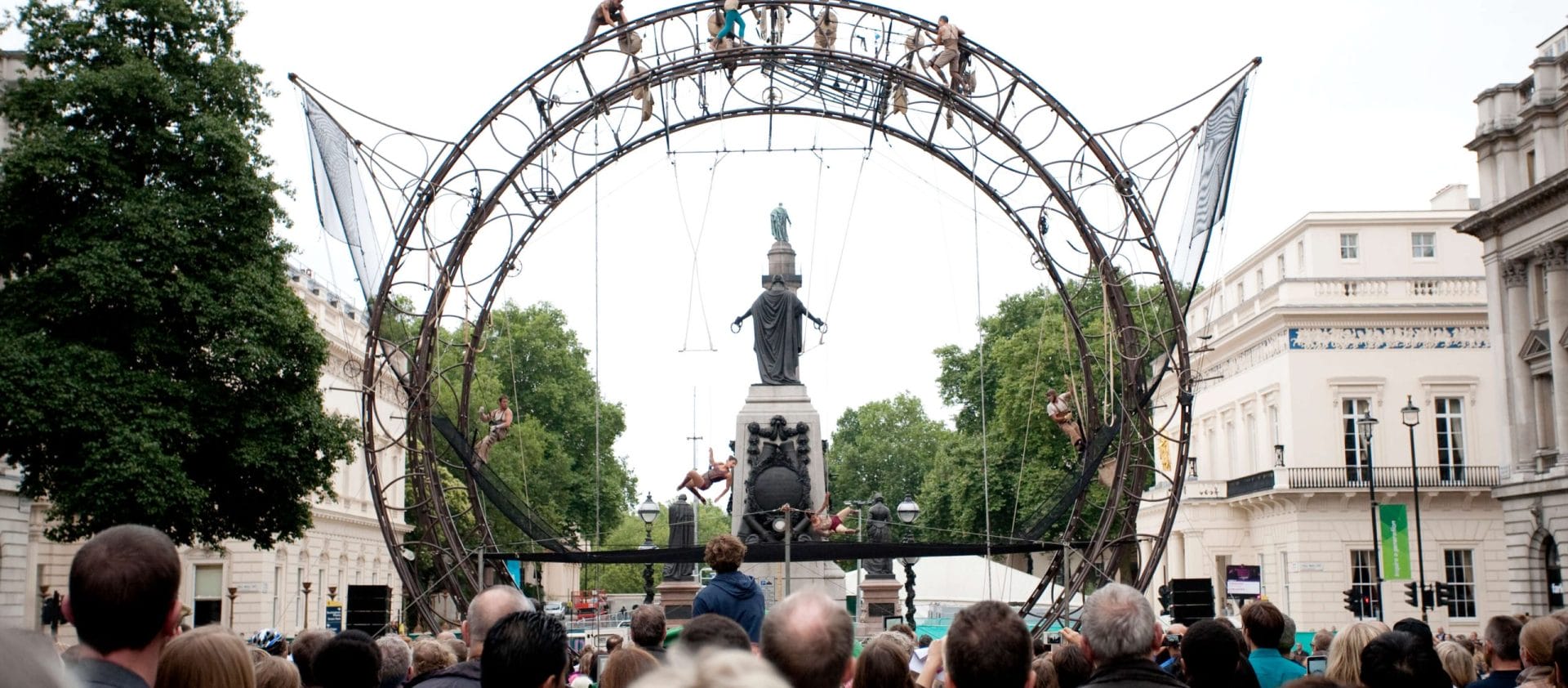an outdoor event featuring acrobats performing on a tall circular wheel-like metal structure, with a large audience watching against a cityscape background
