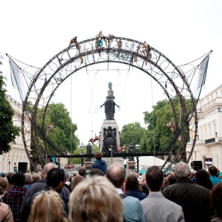 an outdoor event featuring acrobats performing on a tall circular wheel-like metal structure, with a large audience watching against a cityscape background