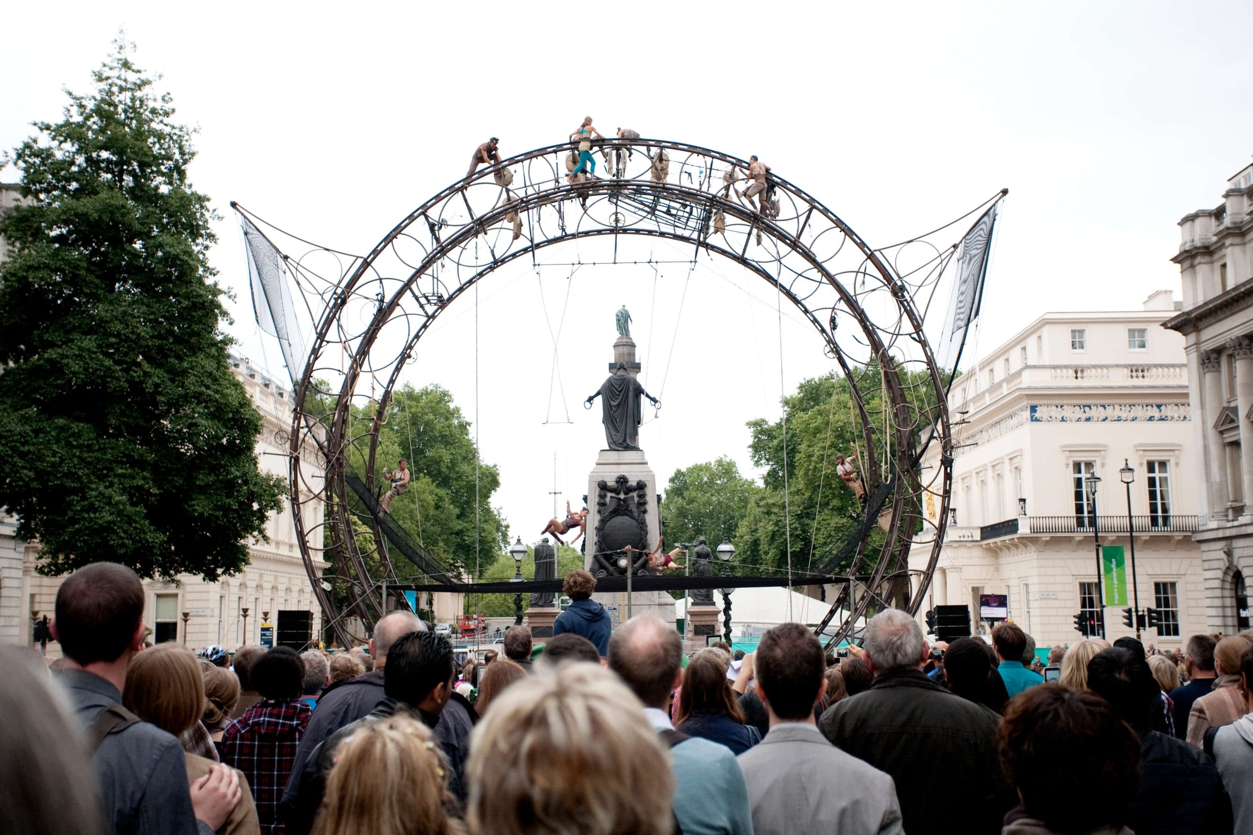 an outdoor event featuring acrobats performing on a tall circular wheel-like metal structure, with a large audience watching against a cityscape background