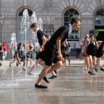 An energetic scene of several jugglers in black costumes running and playing in many water fountains, holding colourful juggling balls in front of Somerset House celebrating juggling in all its facets.