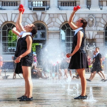 Two performers in coordinated black and white outfits juggle red balls among fountains in front of Somerset House in London.