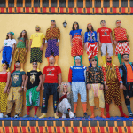 a group of seventeen performers in brightly pattered and colourful, mismatched clothes and all wearing sunglasses form a two-level human tower in front of a yellow wall.