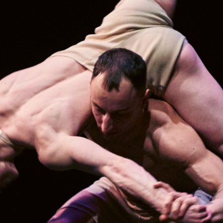 Two dancers on a dark stage, with one dancer lifting the other in a graceful and dynamic lift.
