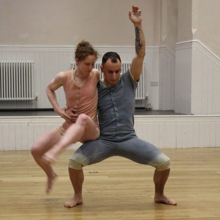 two performers wearing all-in-one buttoned costumes with short sleeves, dance in an indoor rehearsal room, one in blue, with short dark hair holds and supports the other, in pink, with tied up curly red hair