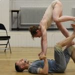 two athletic performers in an indoor rehearsal room one in shorts lies on the floor on their back, the other in shorts and vest is held and climbing on top of them