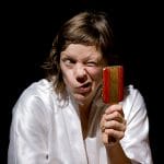 A person with long brown hair wearing a white robe making a playful, exaggerated facial expression while holding up a small mirror, decorated in red and gold, against a dark background