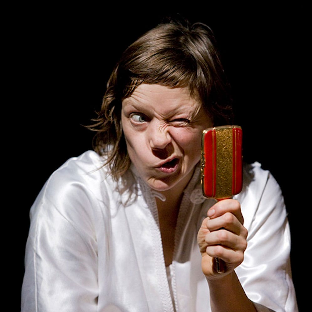 a performer in a silky white shirt, with shoulder length brown hair squints into a small handheld red and gold mirror