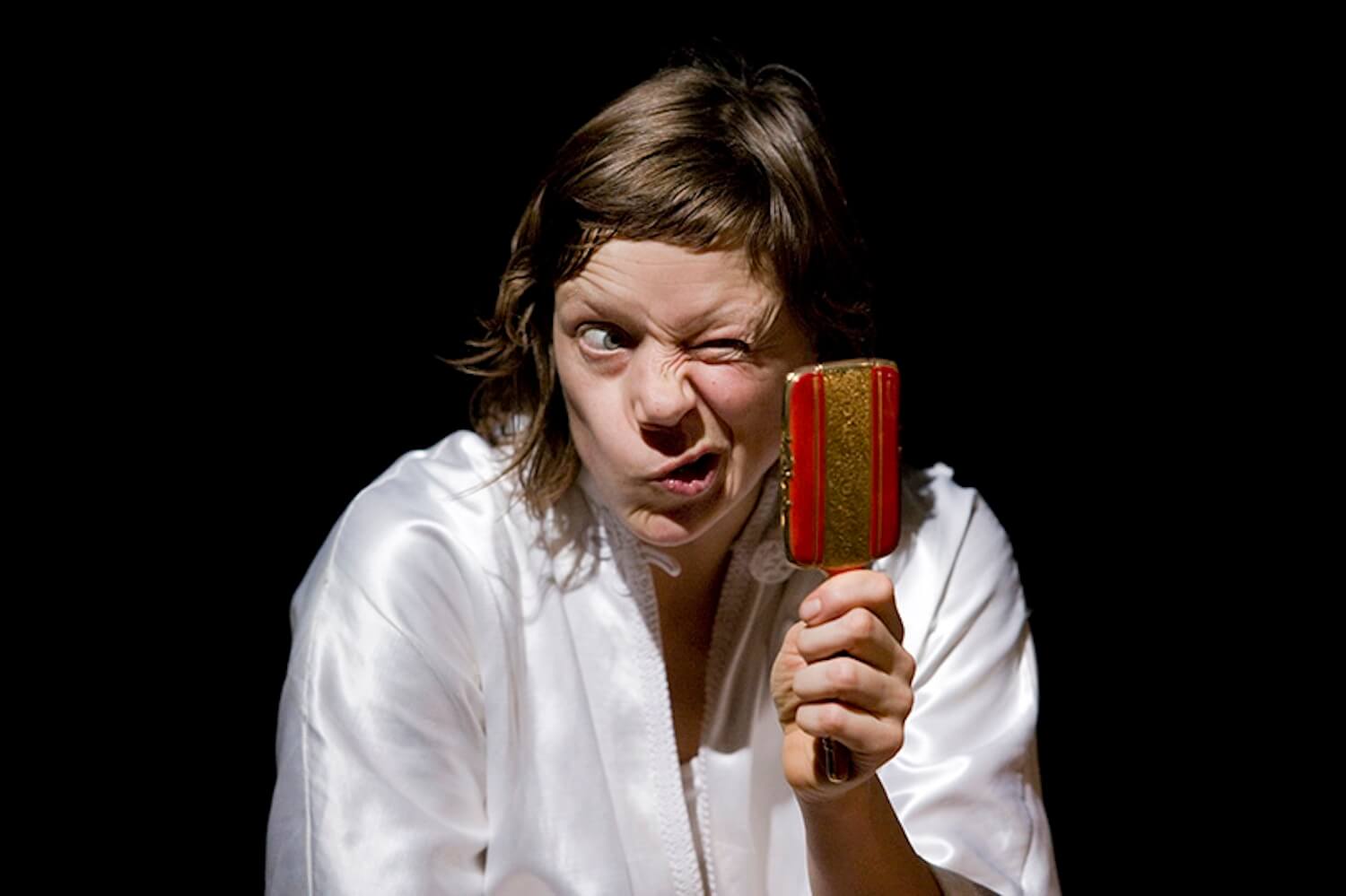 A person with long brown hair wearing a white robe making a playful, exaggerated facial expression while holding up a small mirror, decorated in red and gold, against a dark background