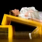 performer wearing a white silky outfit lies on an angled yellow bench looking up to the ceiling