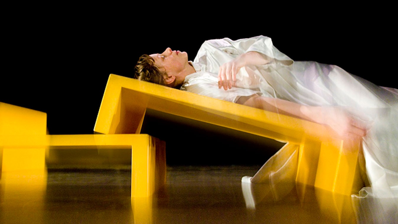 performer wearing a white silky outfit lies on an angled yellow bench looking up to the ceiling