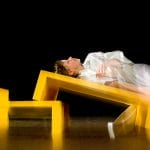 a person lying back on a tilted yellow bench, their body draped in a flowing white robe, with motion shown by blurring adding a sense of dynamic movement against a black background