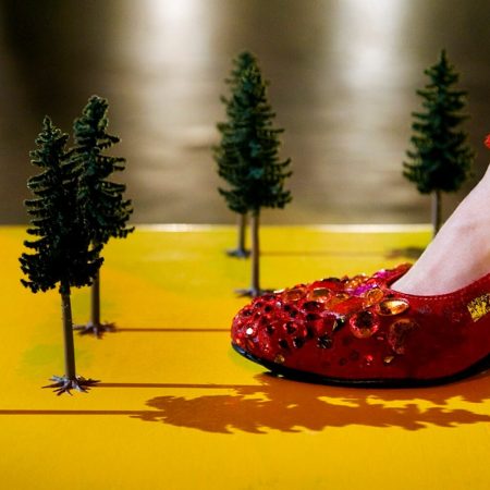 a close-up of a foot in a red shoe decorated with sparkling gems and sequinned embellishments, stepping on a yellow surface surrounded by miniature green fir trees resembling a small, crafted forest scene